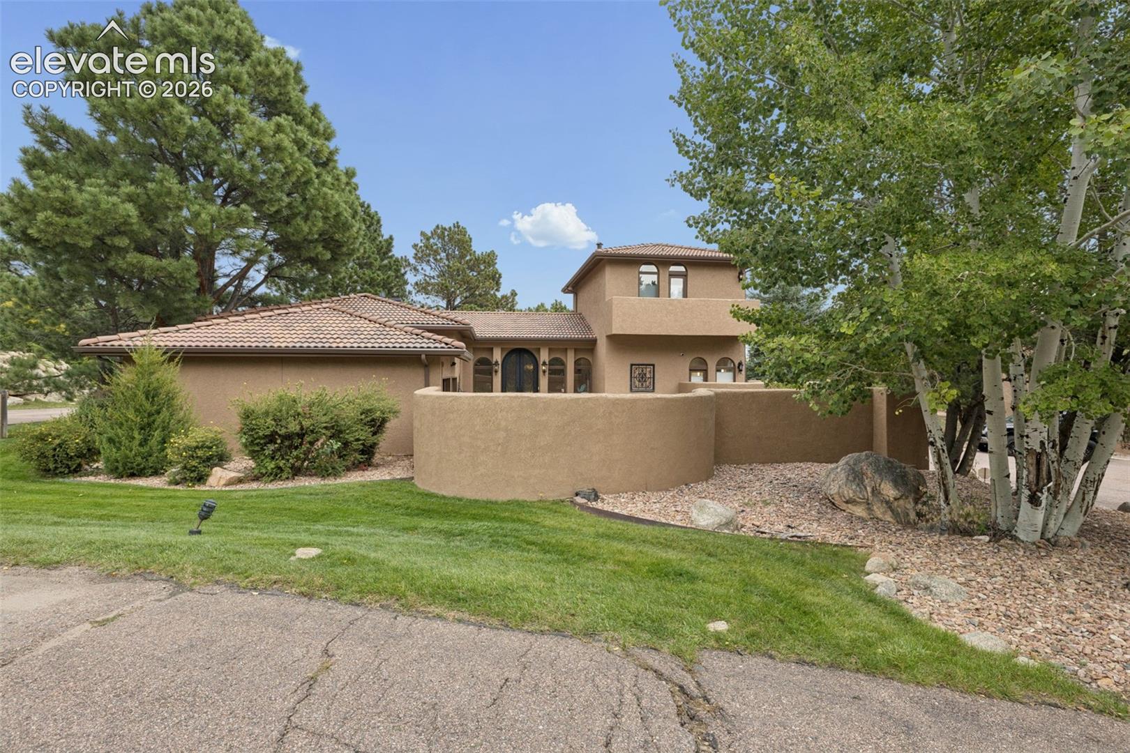 Tan stucco home with a tiled roof, enclosed courtyard wall, lawn, and mature trees.