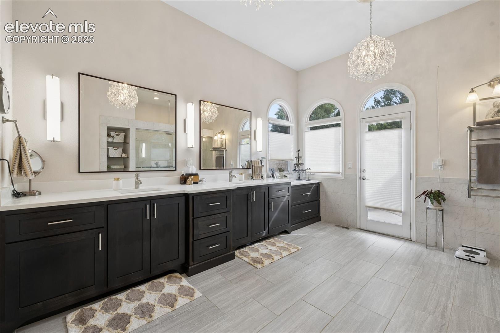 Spacious bathroom with a dark double vanity, arched windows, gray tile floors, and a crystal chandelier.