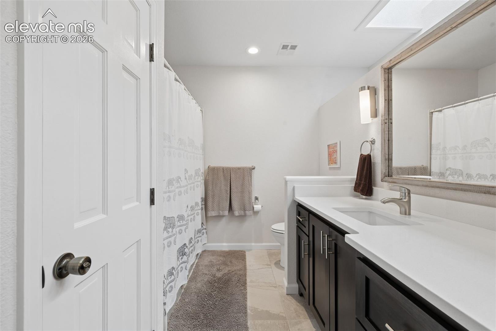 Bathroom with a long white vanity, dark cabinets, elephant-patterned shower curtain, and ceiling skylight.