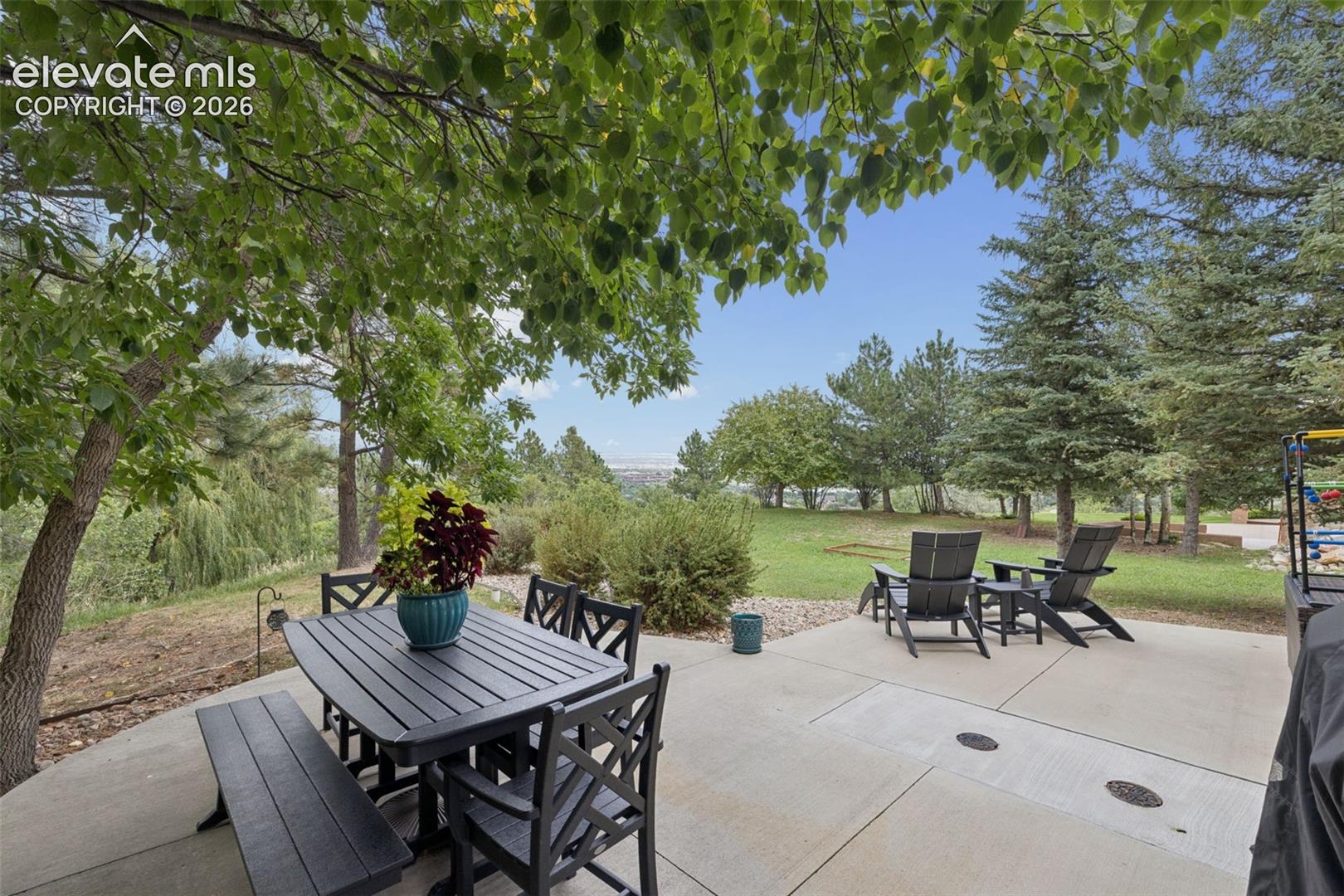 Furnished patio with dining and lounge seating beneath trees, overlooking a lawn and distant city view.