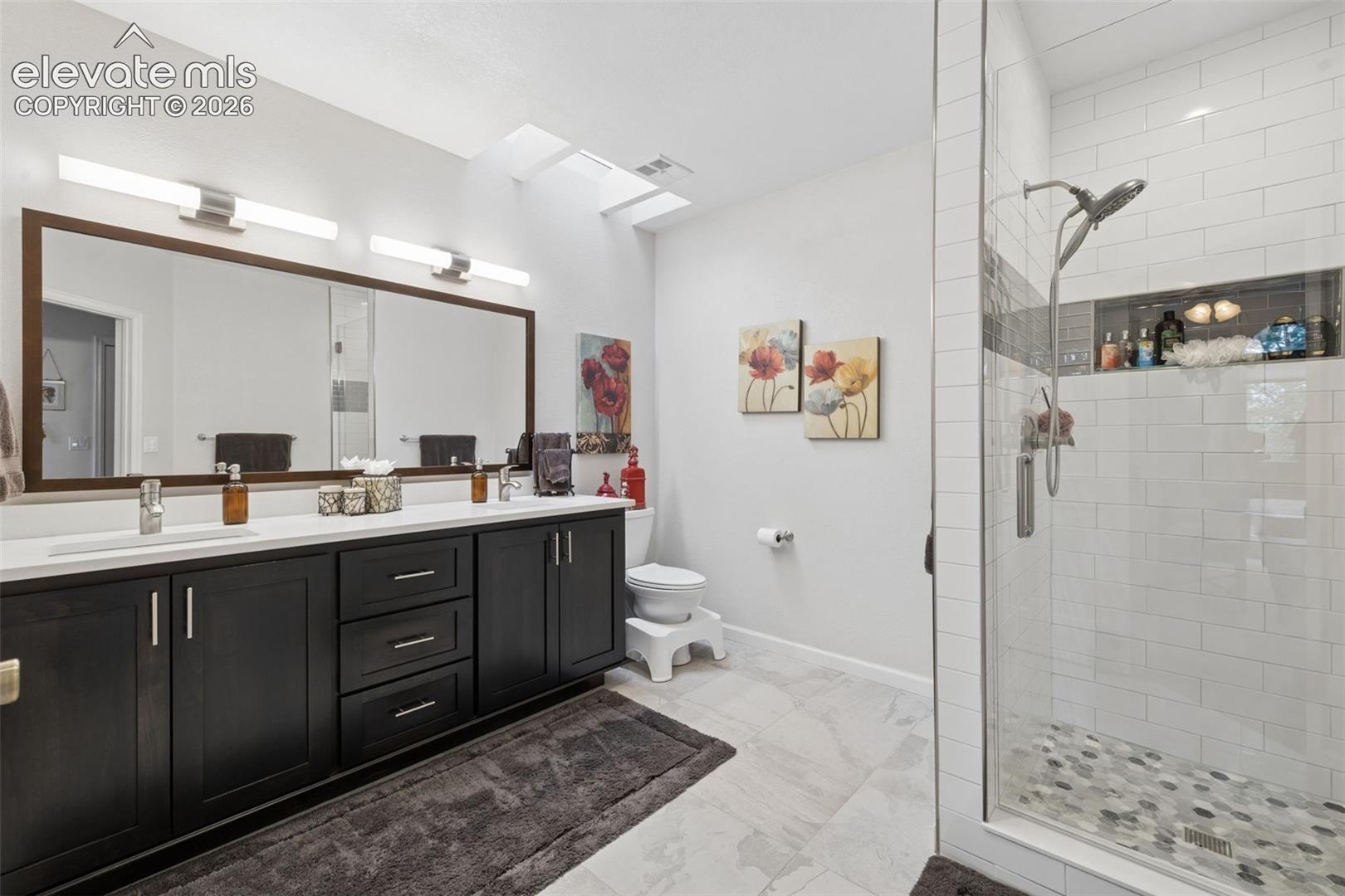 Bathroom with a dark double vanity, skylights, and a glass-enclosed subway-tile shower.