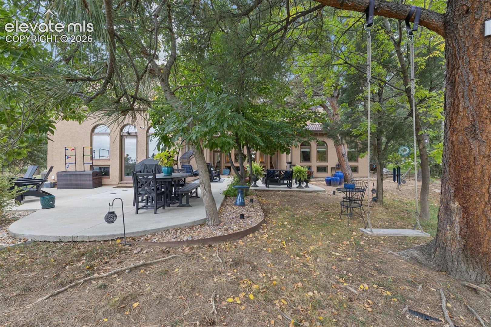 Wooded backyard patio with outdoor dining furniture and a swing beneath a large tree.