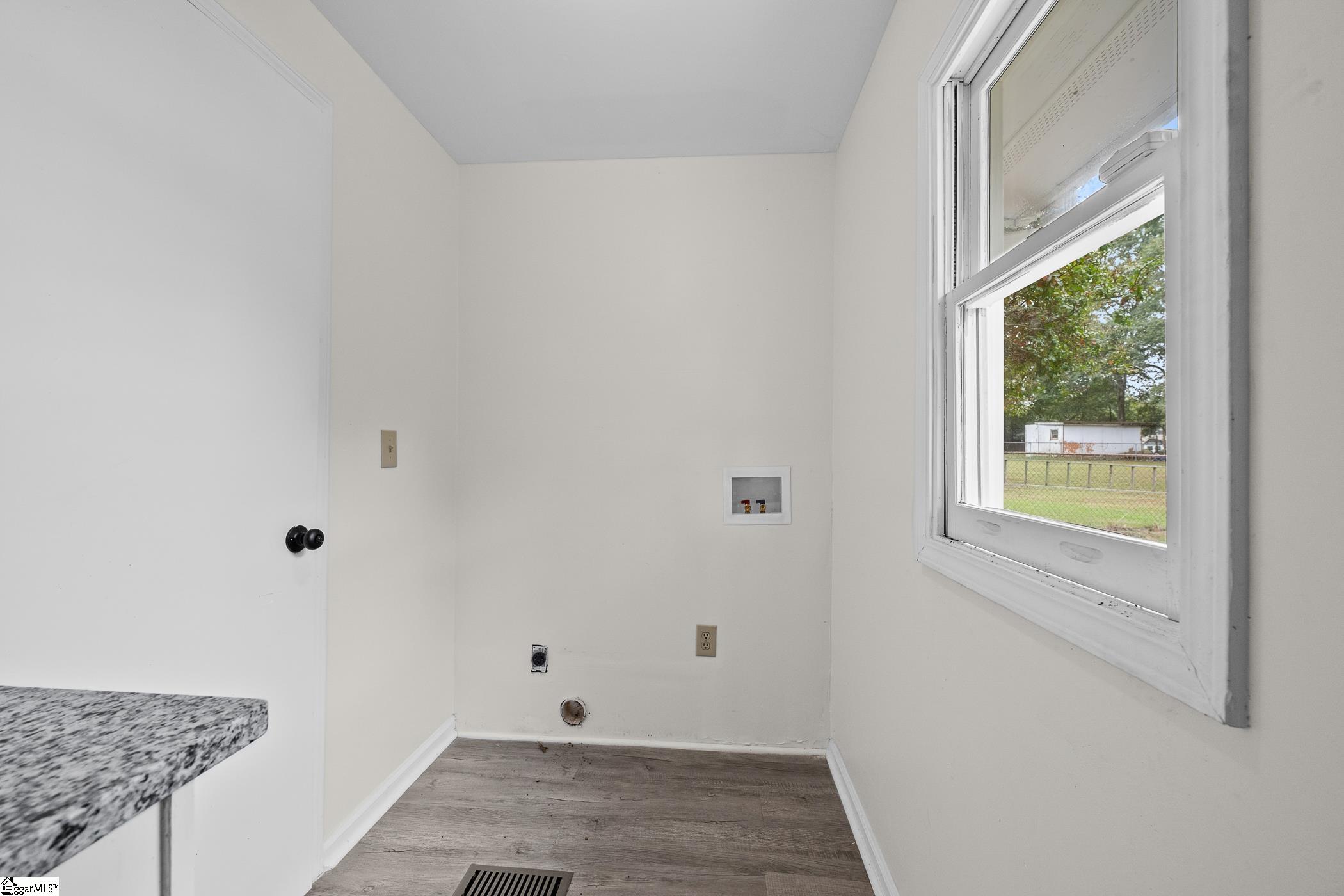 Empty laundry room with washer and dryer hookups, gray flooring, and a large window overlooking a fenced yard.