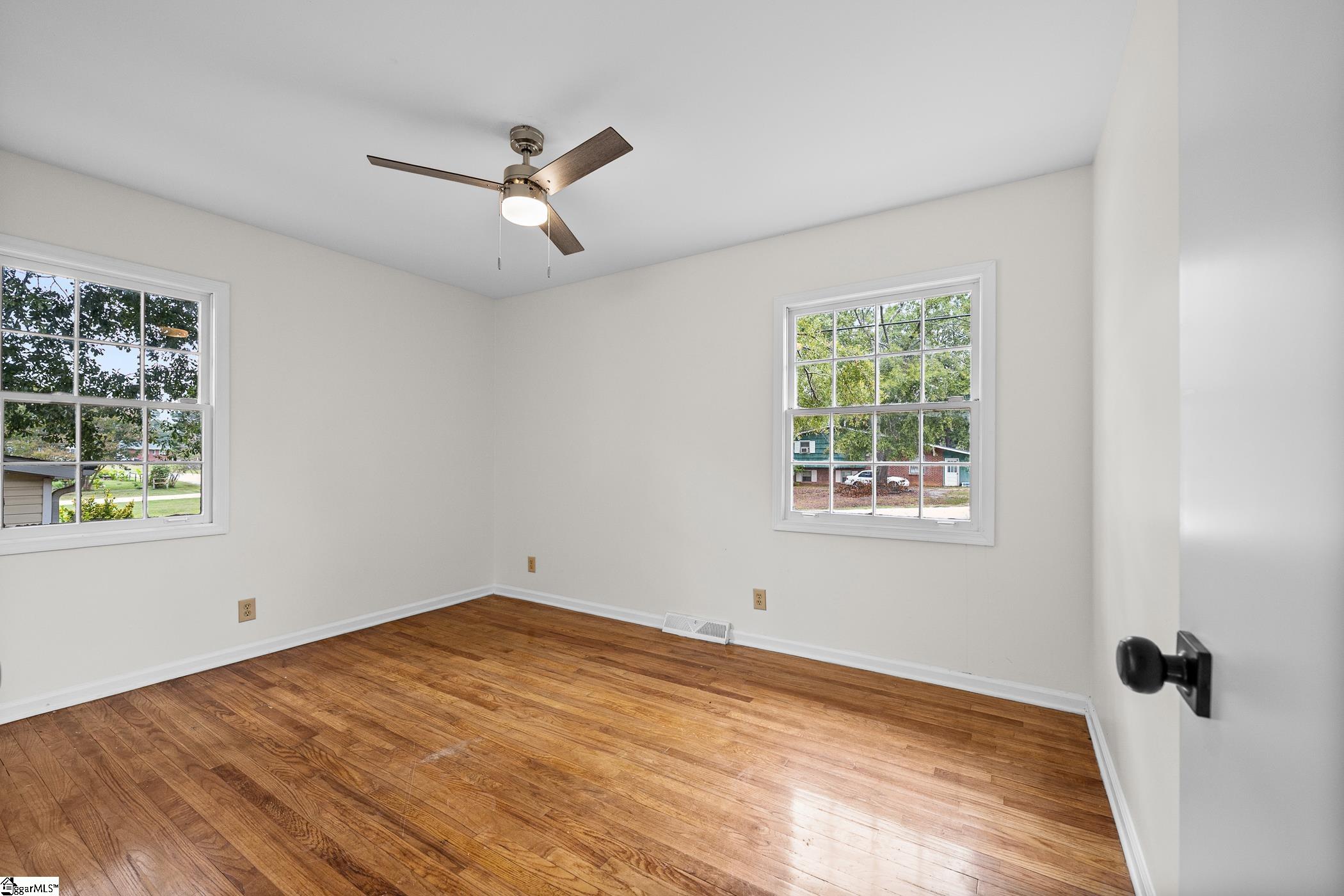 Empty room with polished hardwood floors, two windows, and a ceiling fan.