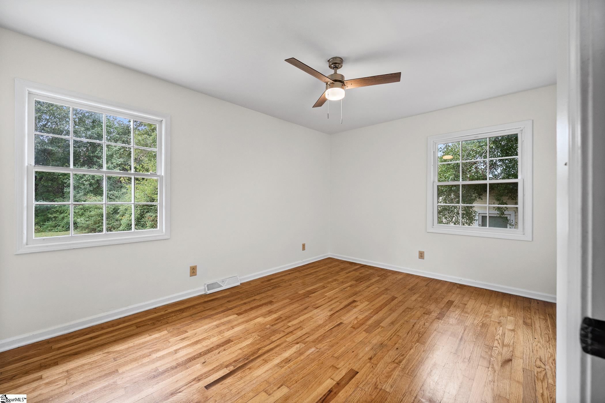 Empty room with hardwood floors, two windows, and a wood ceiling fan.