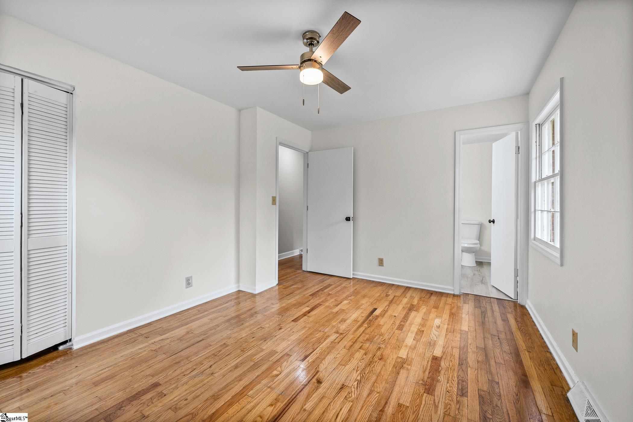 Empty bedroom with hardwood floors, ceiling fan, and open doorway to a bathroom.