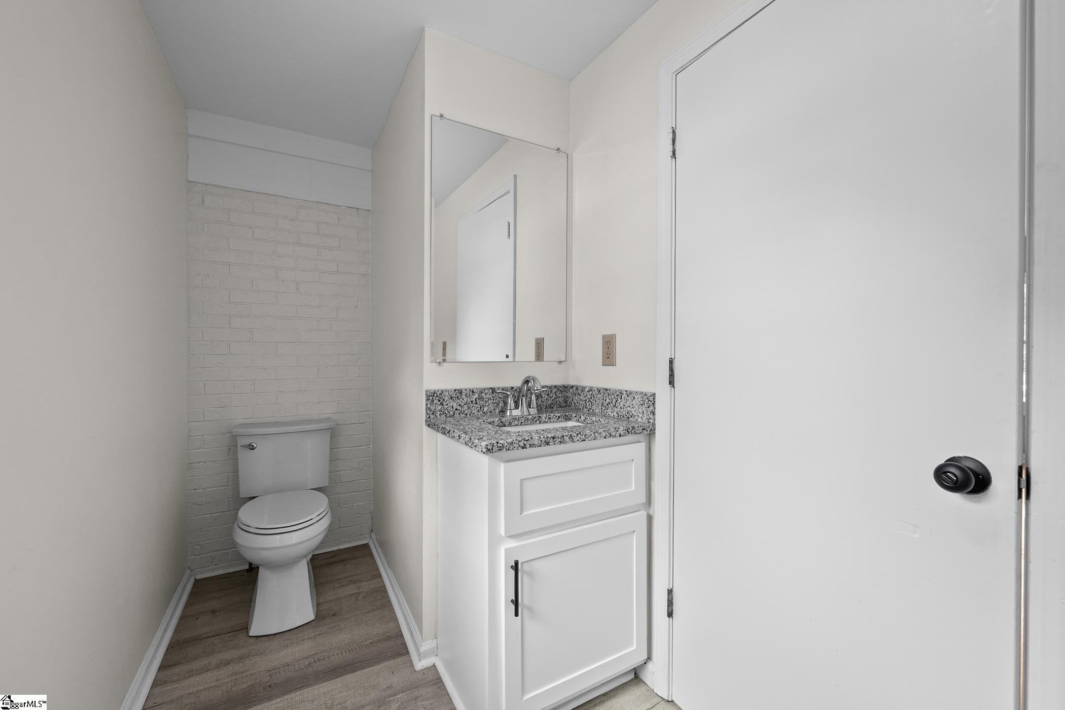 Bathroom with a granite-topped vanity, wall mirror, and toilet against a painted brick wall.