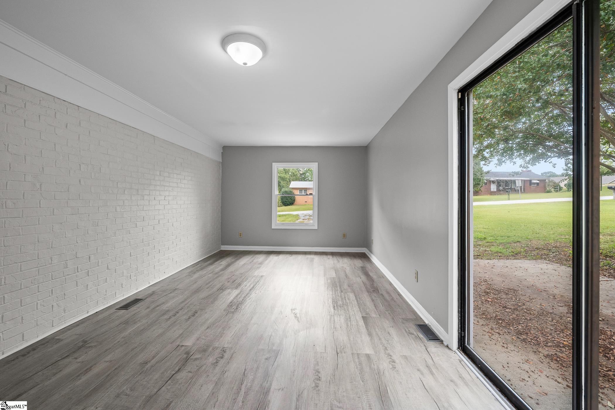 Empty room with a white-painted brick wall, gray plank flooring, and a sliding glass door to the yard.