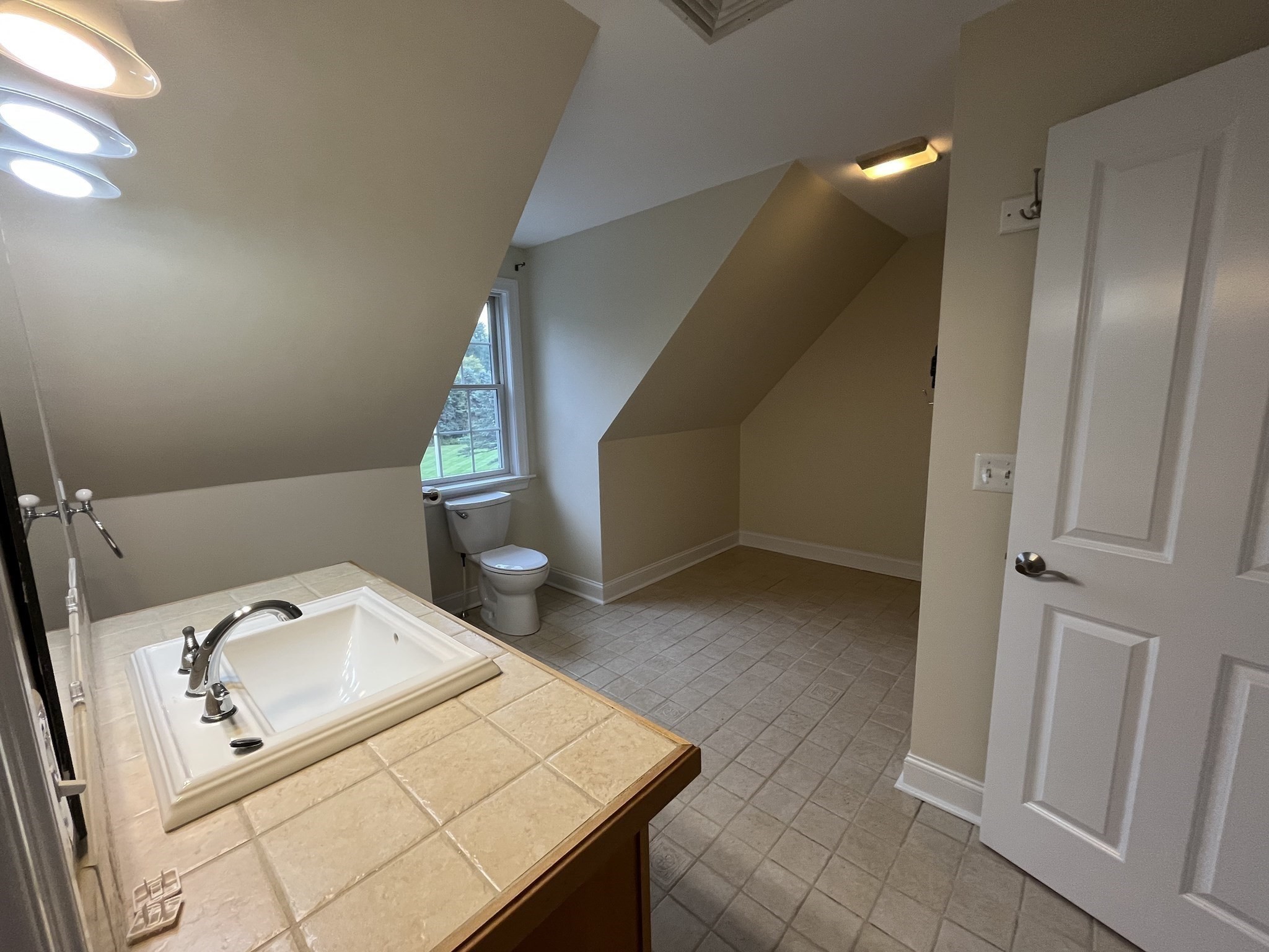 Attic bathroom with sloped ceilings, tiled vanity, white sink, and toilet beside a window.