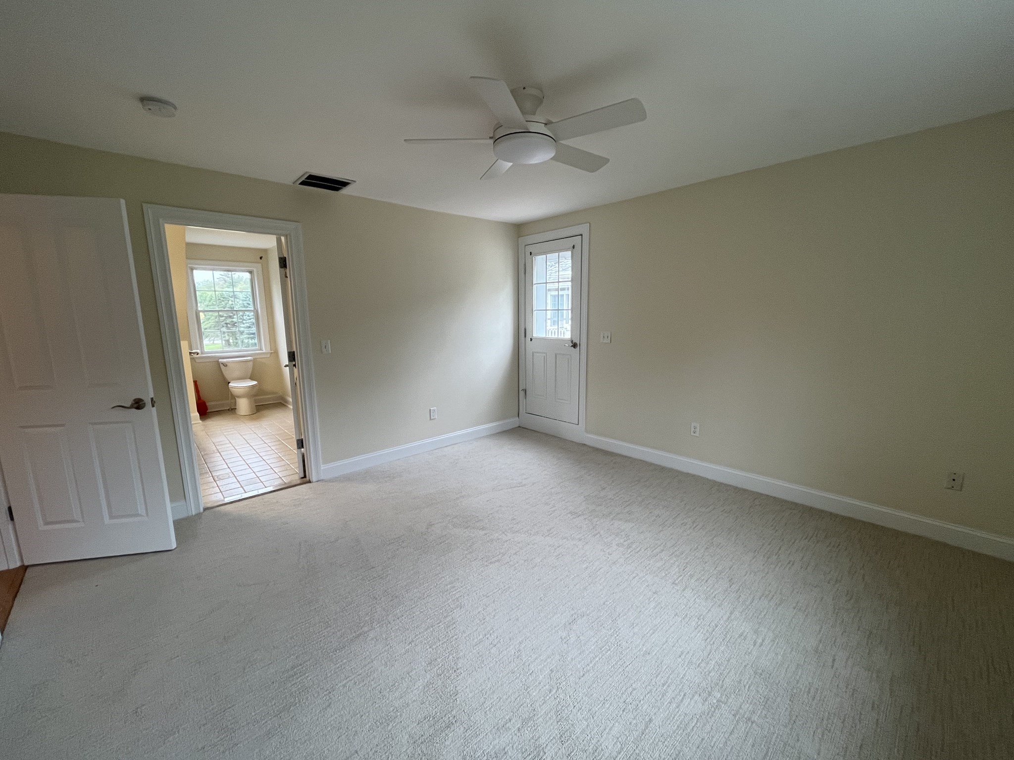 Empty carpeted room with a ceiling fan and an open doorway to a tiled bathroom.