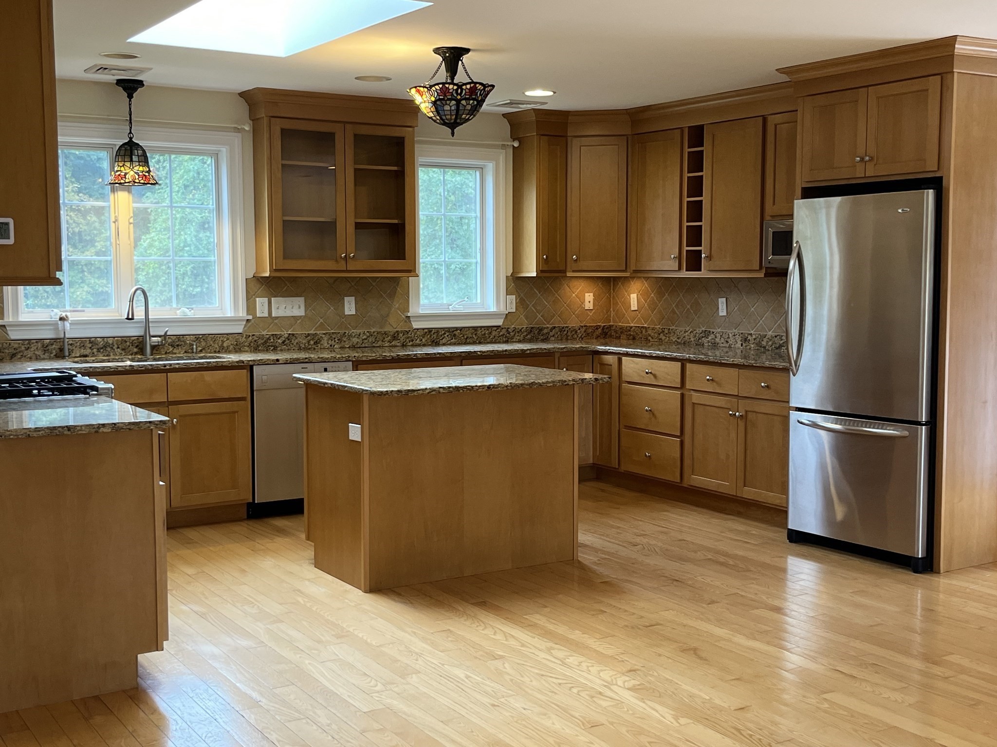 Kitchen with warm wood cabinetry, granite counters, central island, stainless steel refrigerator, and skylight.