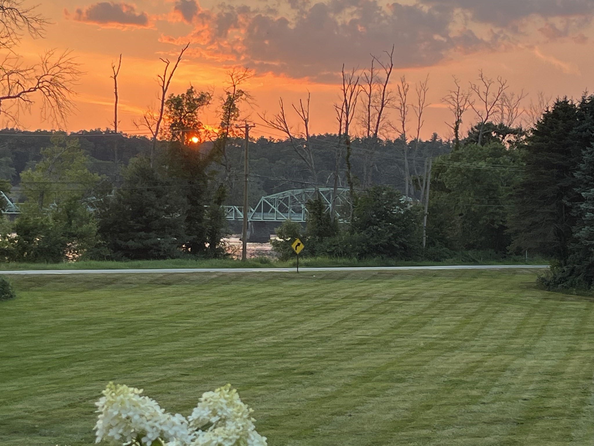 Mowed lawn overlooking a wooded river and truss bridge beneath an orange sunset.