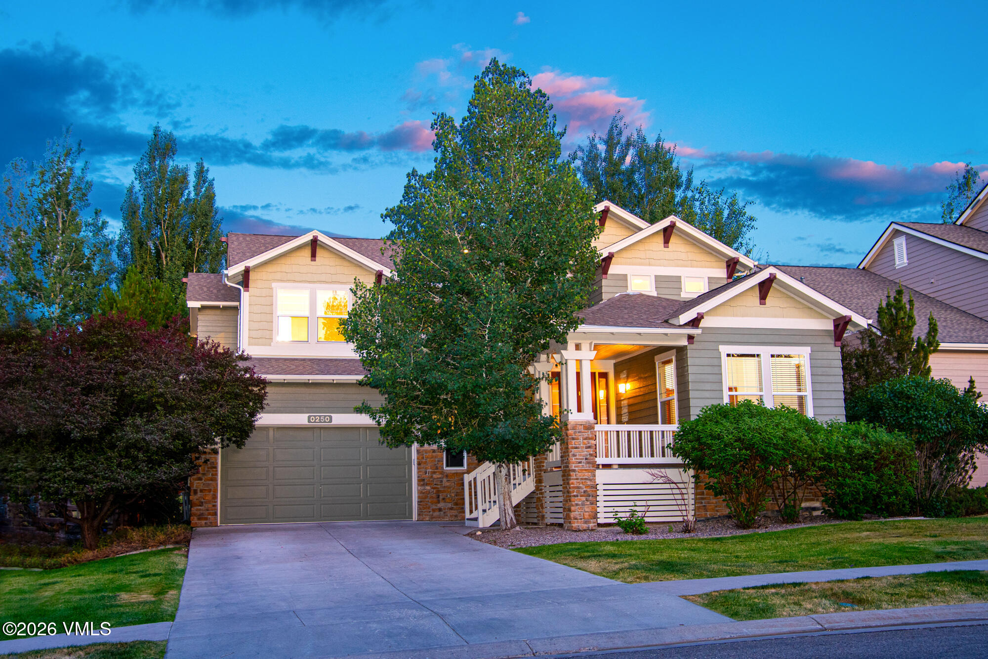 Two-story gray and cream house with stone accents, front porch, garage, and landscaped yard at dusk.