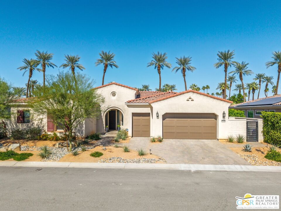 White stucco home with an arched entry, tan garage doors, desert landscaping, and tall palms.