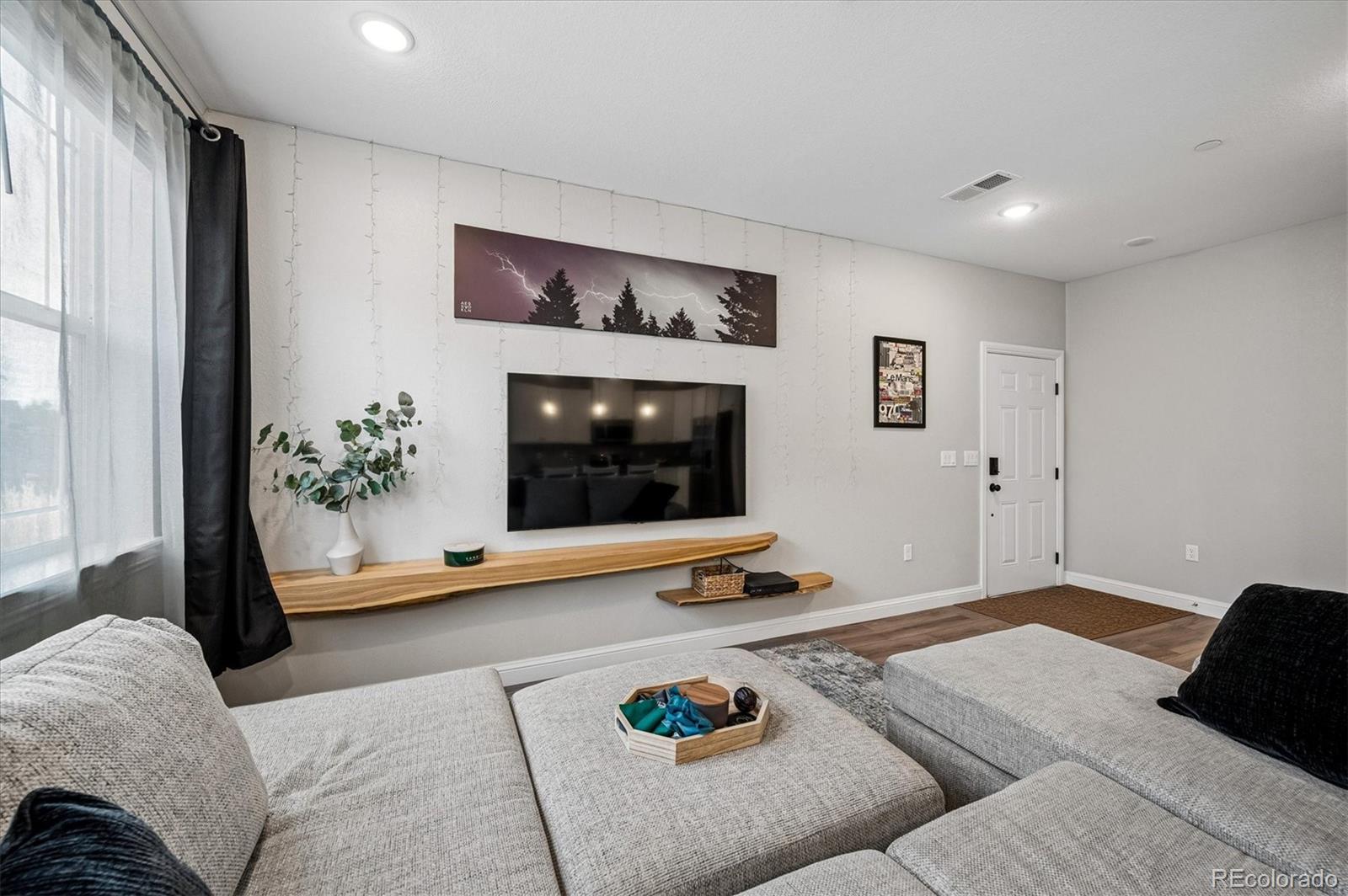 Furnished living room with a gray sectional, wall-mounted television, and live-edge wood shelf.