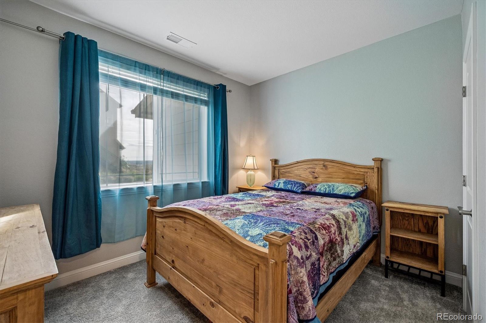 Bedroom with a wooden bed, colorful quilt, gray carpet, and teal curtains framing a large window.