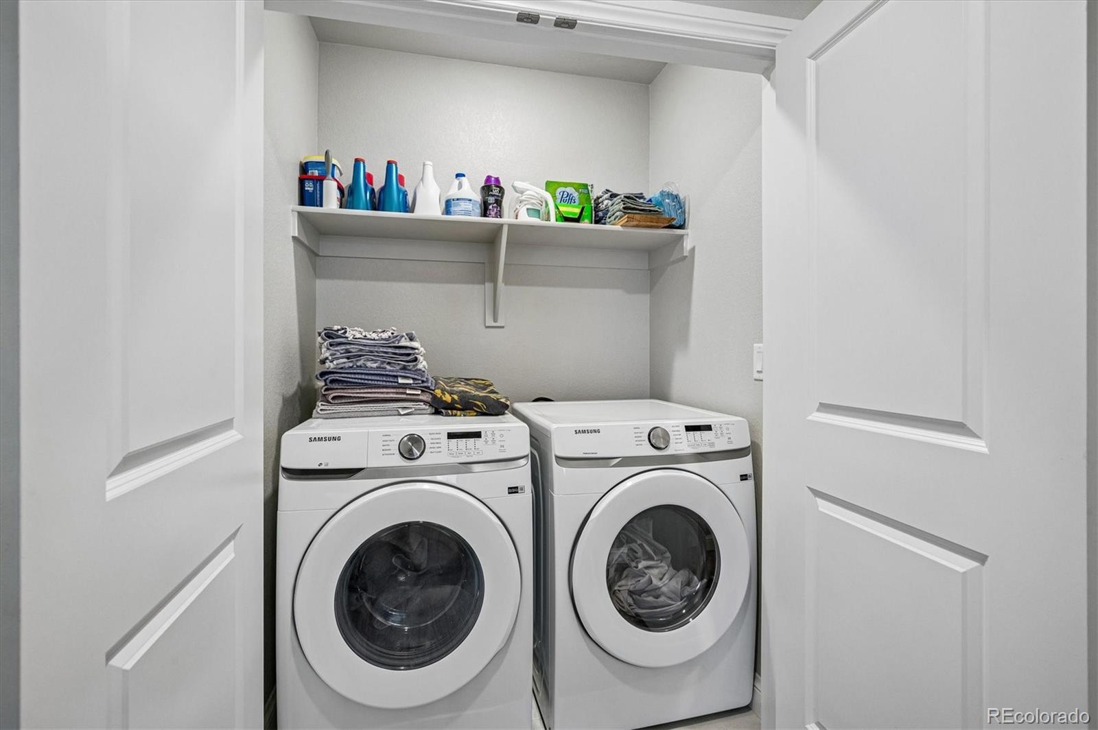 Laundry room with side-by-side white front-loading washer and dryer beneath a shelf of supplies.