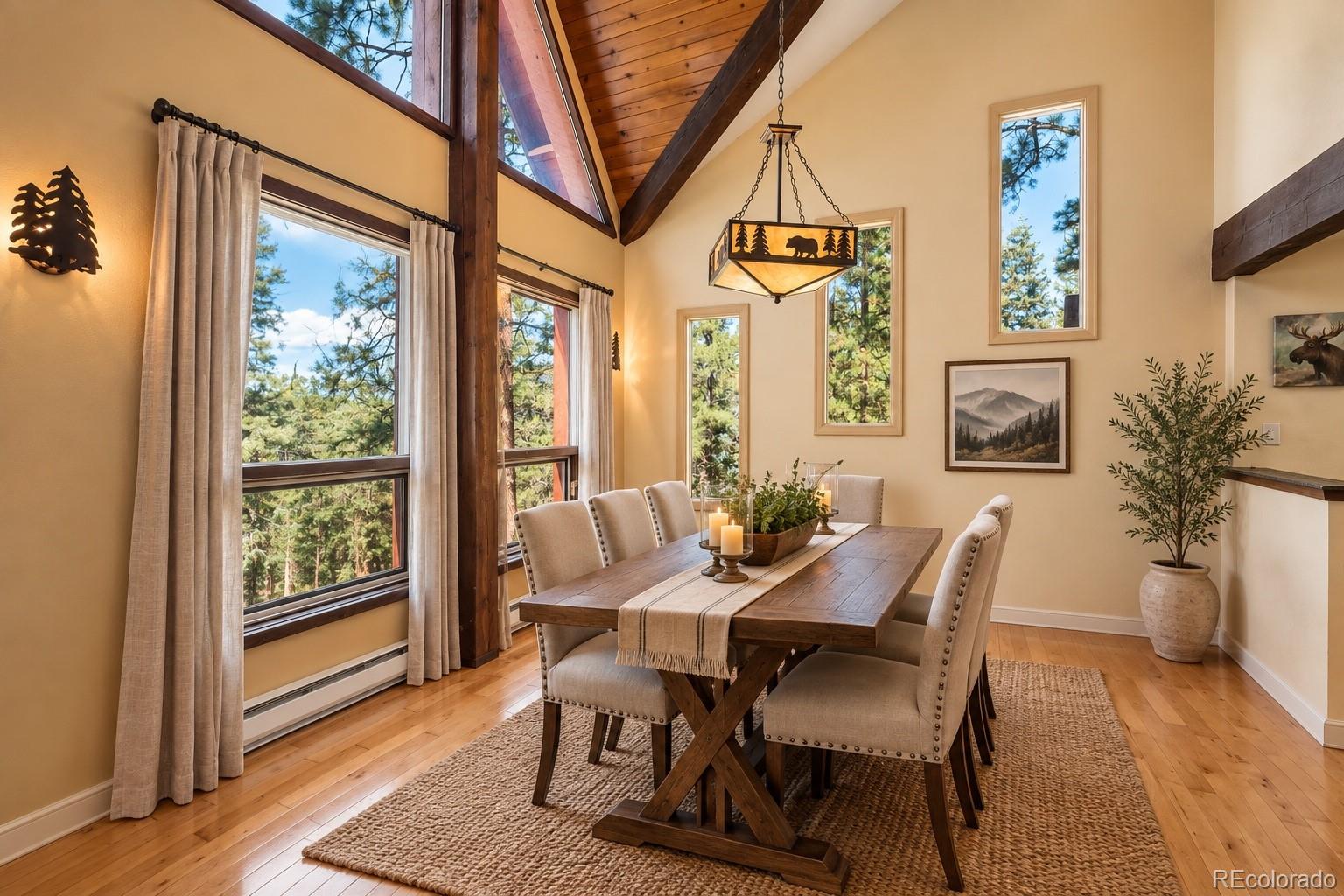 Dining room with a rustic wood table, vaulted ceiling, and tall windows overlooking pine trees.