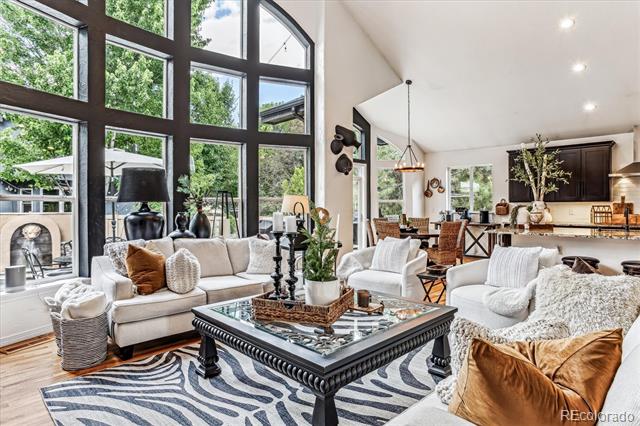 Open-plan living room with tall black-framed windows, white sofas, and a view of leafy greenery.