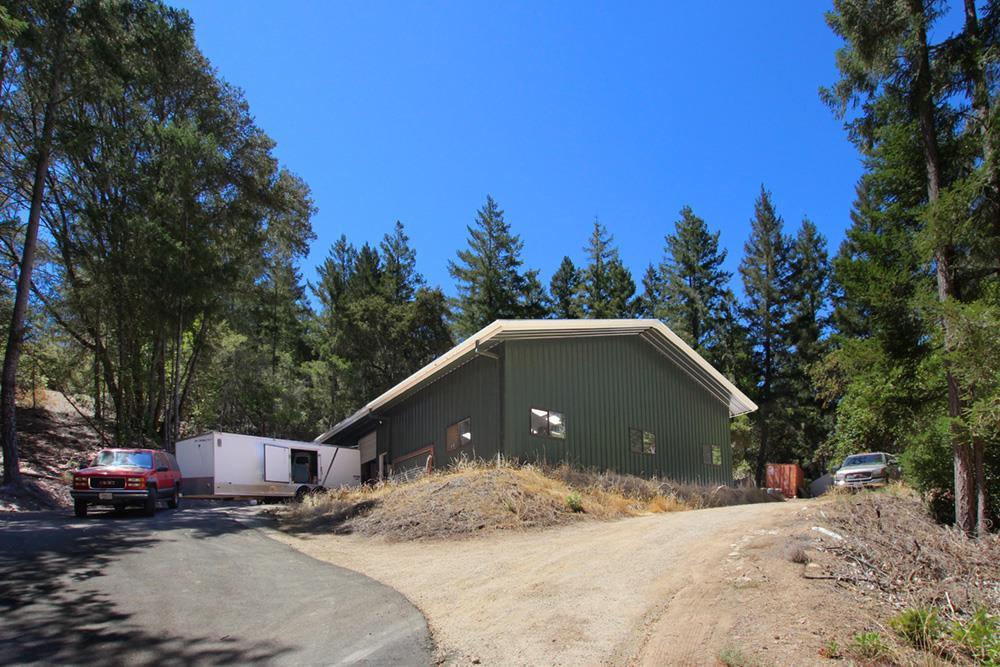 Green metal-sided building among tall trees, with a dirt driveway and vehicles nearby.