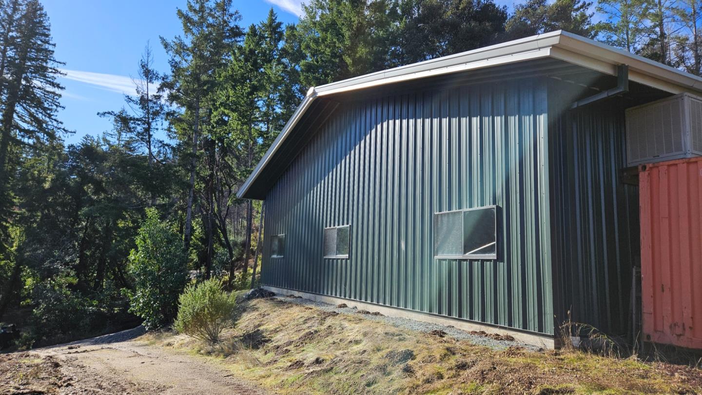 Green metal-sided building beside a gravel path, with a red shipping container and wooded surroundings.