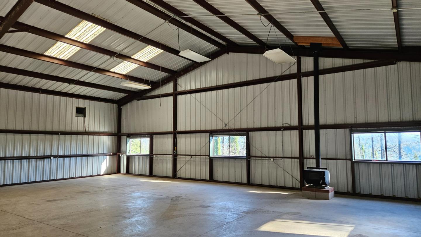 Spacious metal workshop with exposed steel beams, concrete floor, windows, and a wood-burning stove.