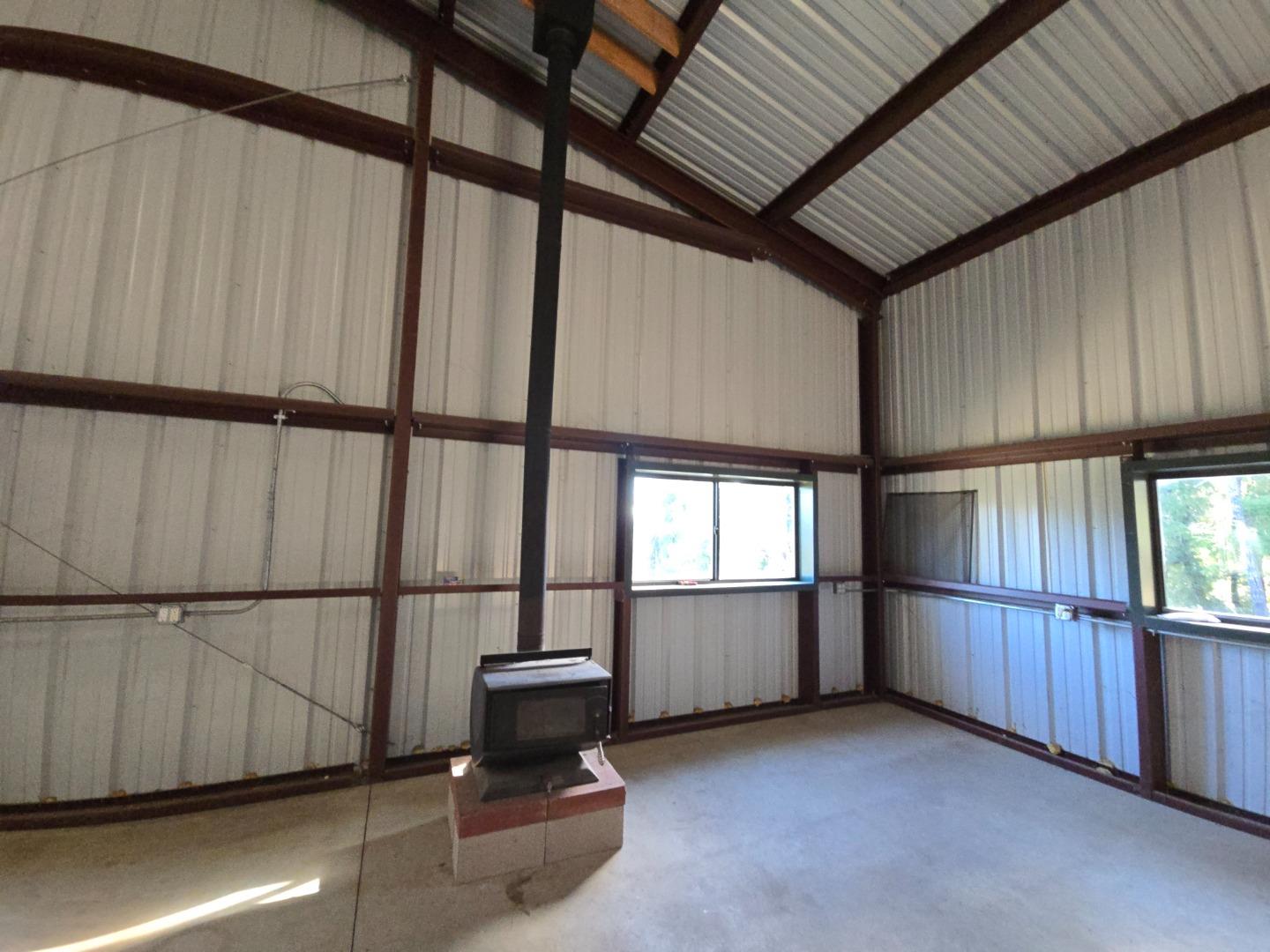 Vacant metal workshop with exposed steel framing, windows, and a freestanding wood stove.