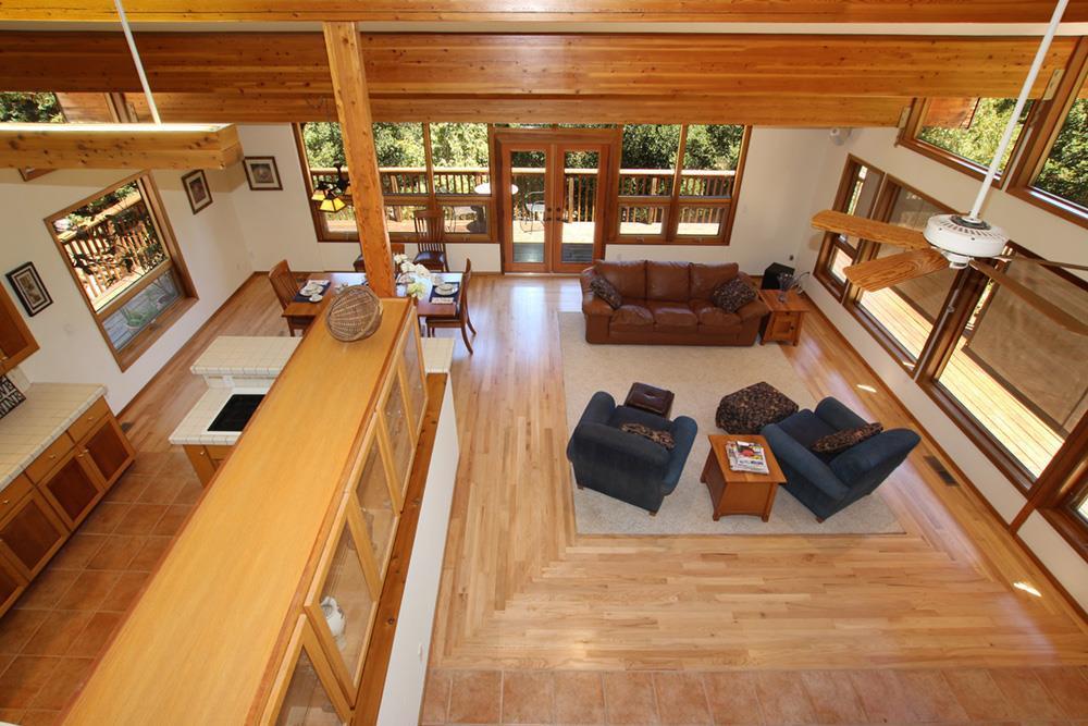 Open-plan living room with exposed wood beams, hardwood floors, and large windows overlooking a deck.