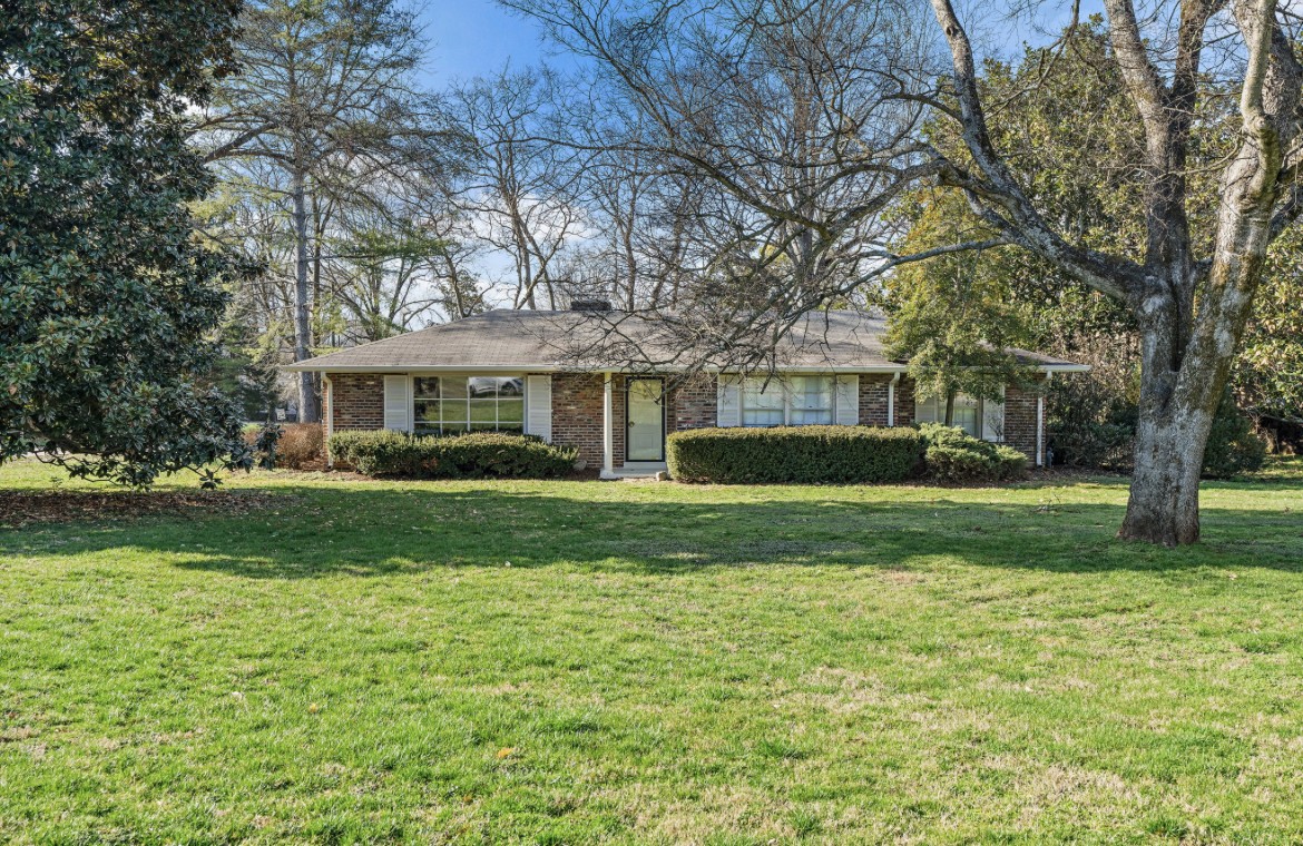 Single-story brick house behind a broad lawn, framed by mature trees.