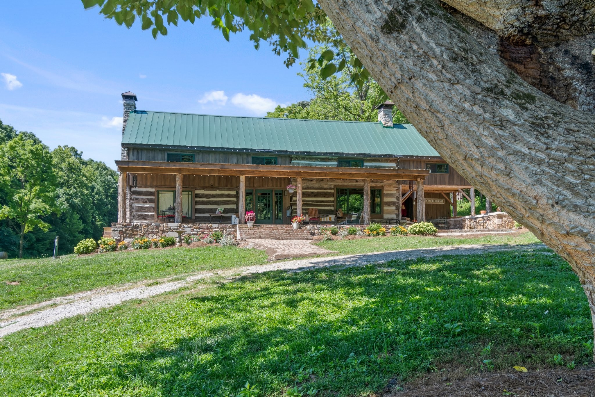 Rustic log cabin with a green metal roof, broad porch, and flower beds beneath a large tree.