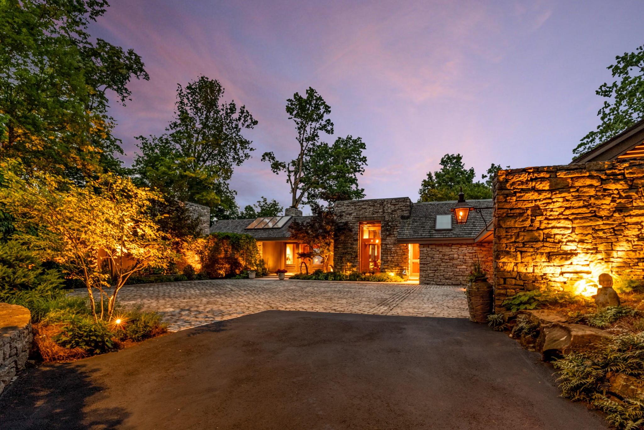 Stone house with a broad paved driveway and warmly lit landscaping at dusk.