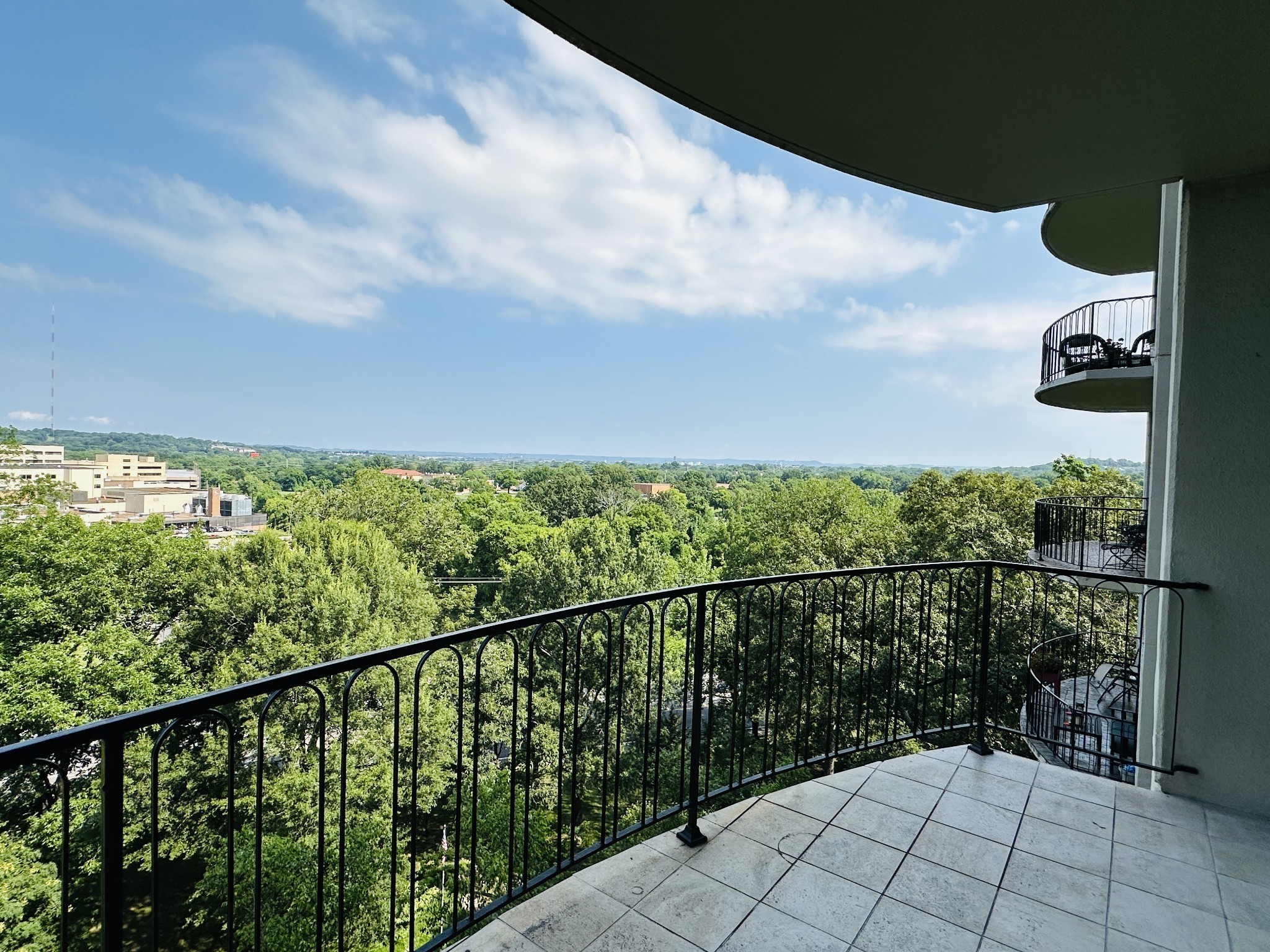 Tiled apartment balcony overlooking green trees and distant buildings, with black metal railings.