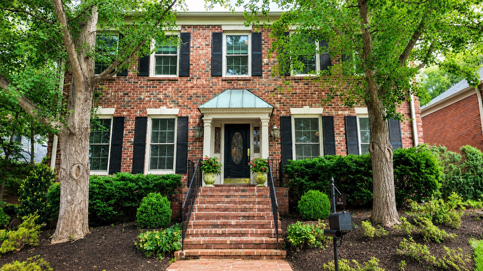 Two-story brick house with black shutters, brick entry steps, and mature trees framing the front.