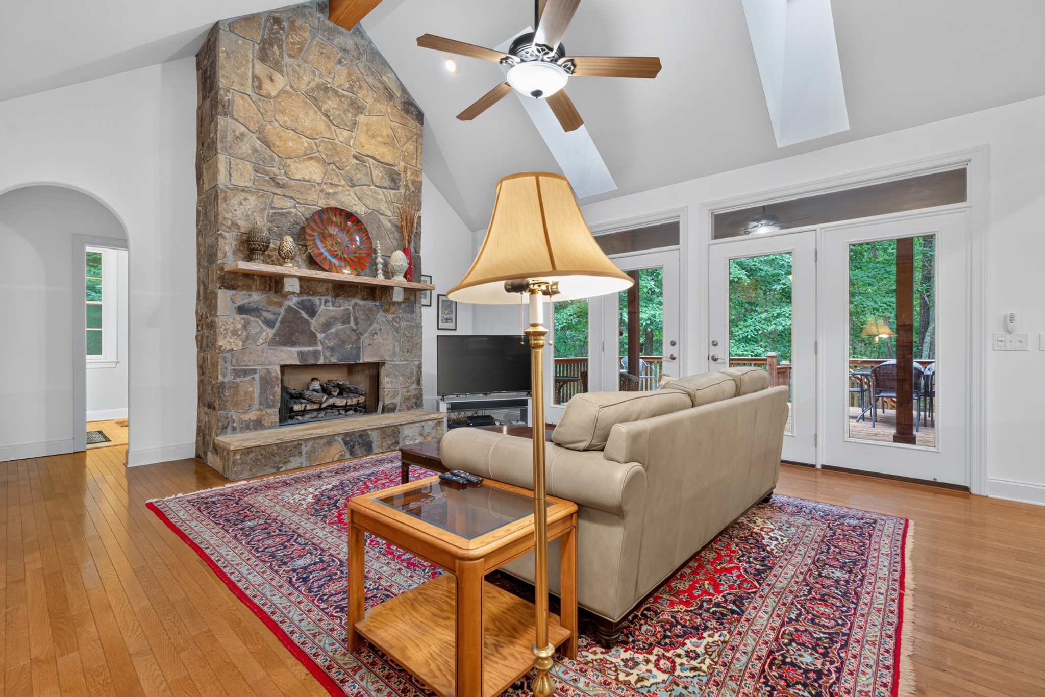 Living room with a tall stone fireplace, vaulted ceiling, beige sofa, and glass doors to a wooded deck.
