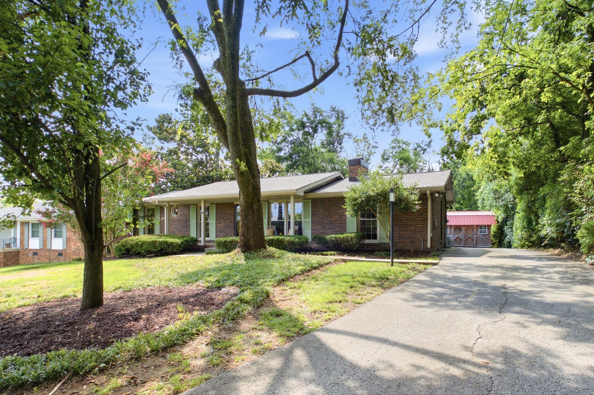 Brick ranch-style house shaded by mature trees, with a broad driveway leading to a red-roofed outbuilding.