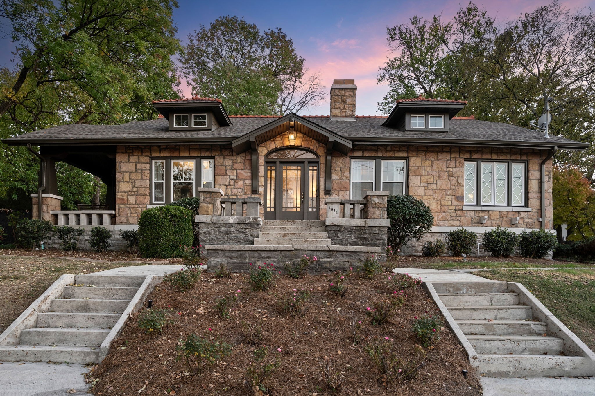 Stone house with a central arched entrance, broad front steps, and garden beds at dusk.
