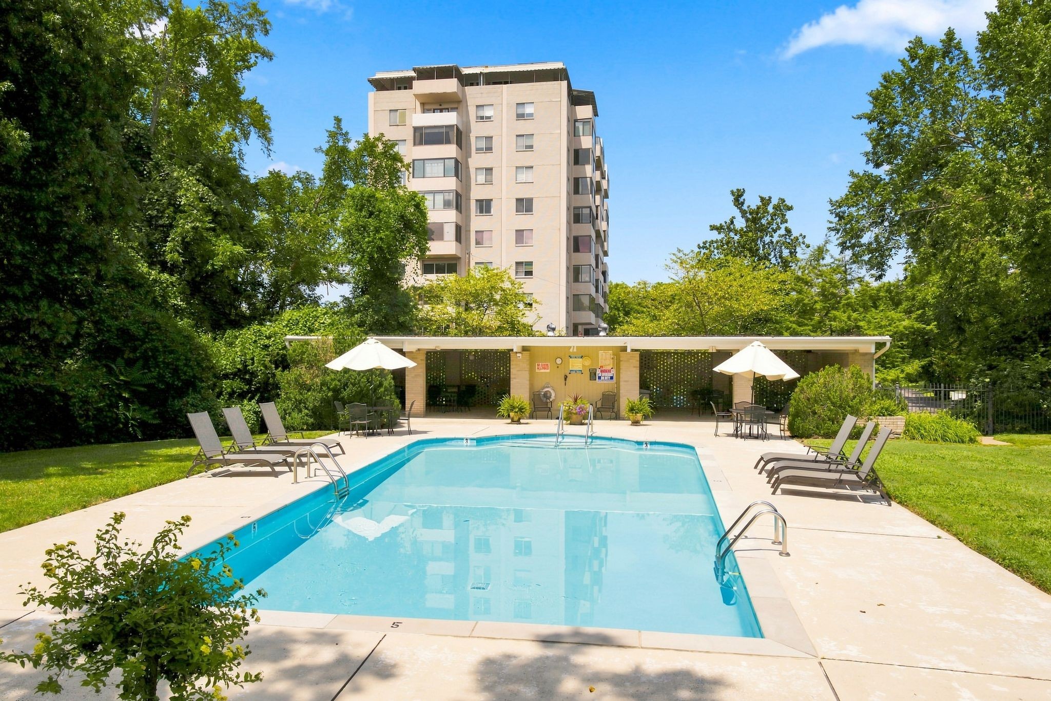 Outdoor pool with lounge chairs and umbrellas in front of a tree-surrounded apartment building.