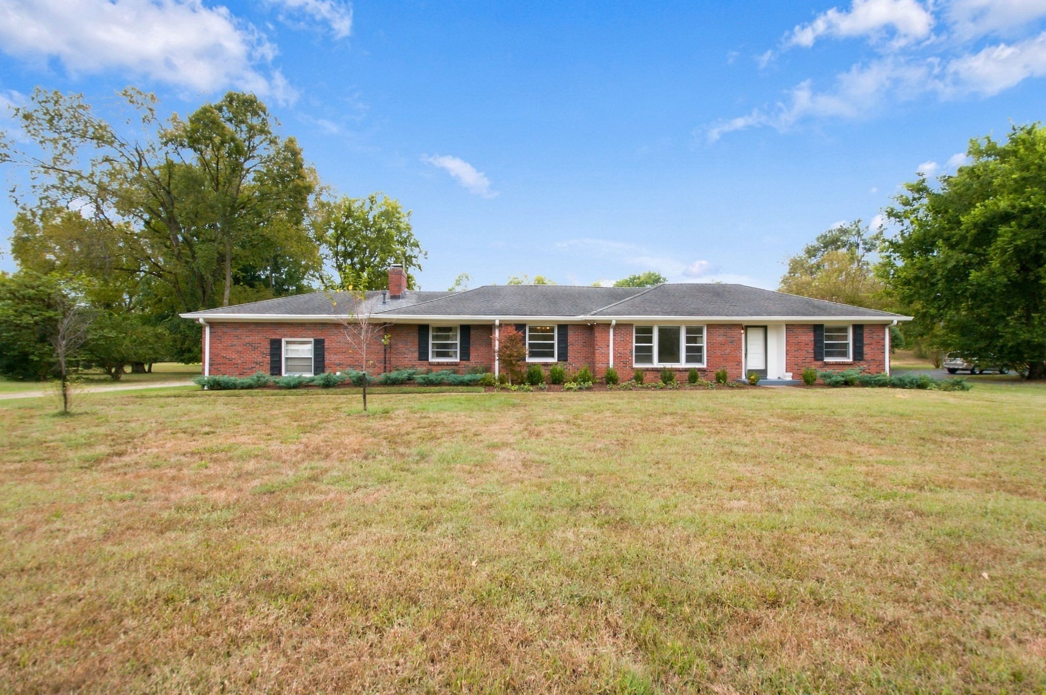 Single-story red-brick ranch house with dark shutters and a broad lawn framed by mature trees.
