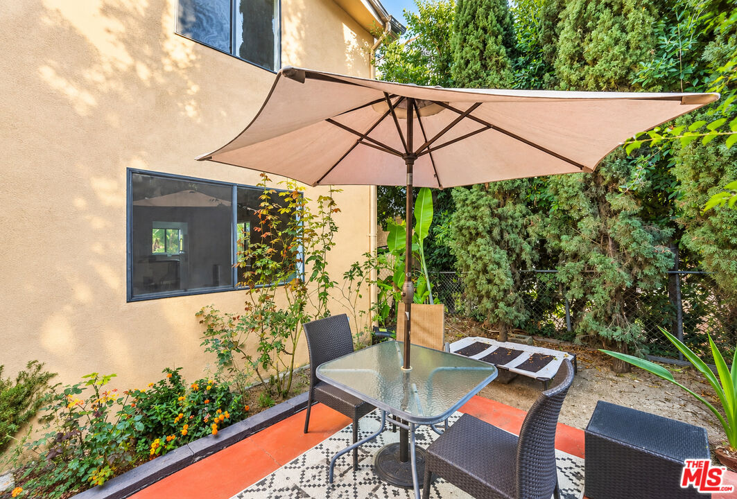 Outdoor patio with a glass-top dining table, large umbrella, patterned rug, and lush greenery.