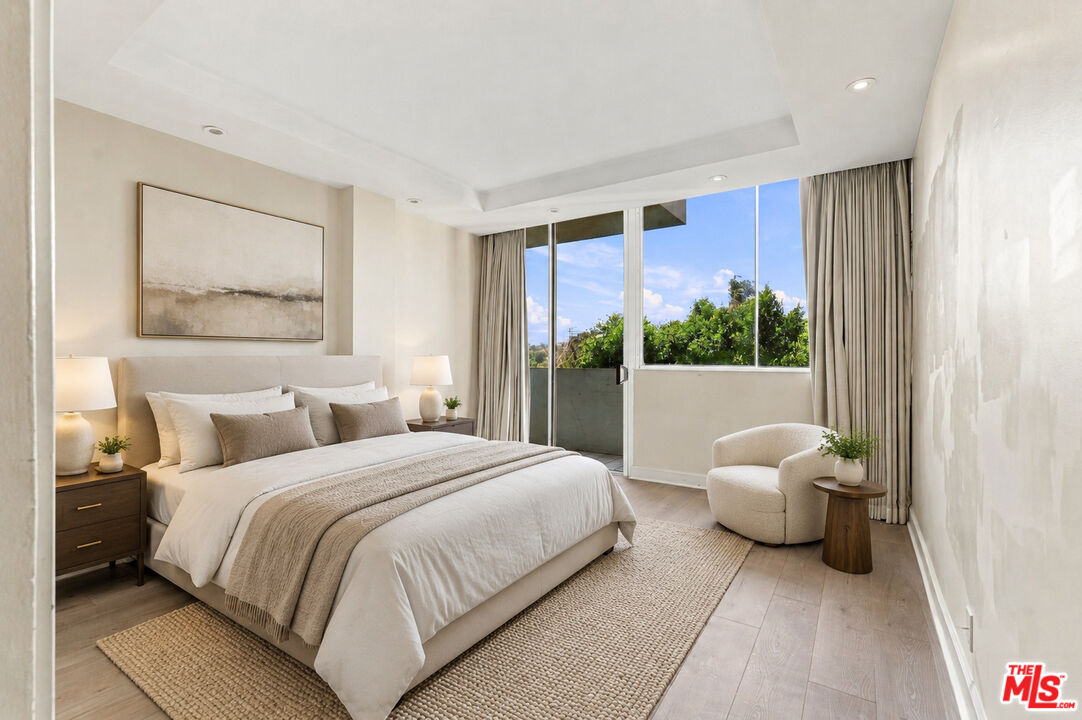 Bedroom with a neutral upholstered bed, abstract wall art, and windows overlooking leafy trees.