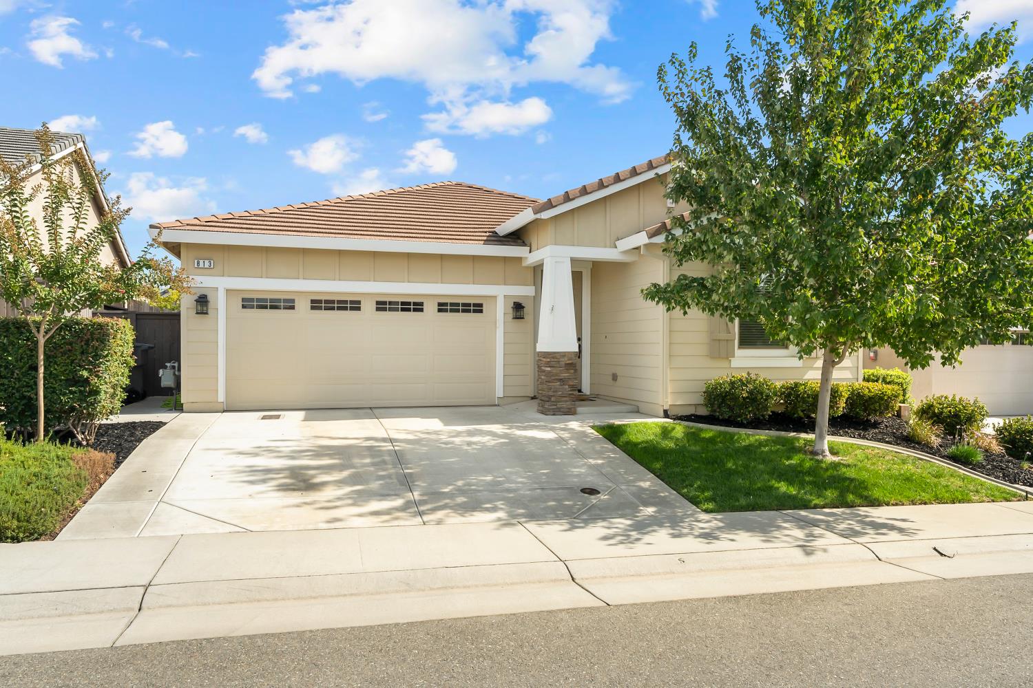 Beige single-story home with a two-car garage, broad driveway, and leafy tree in the front yard.