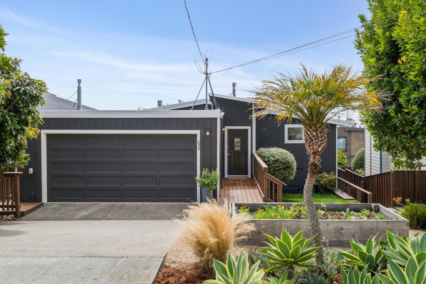 Charcoal-gray home with a wide garage, wooden entry walkway, palm tree, and landscaped front garden.