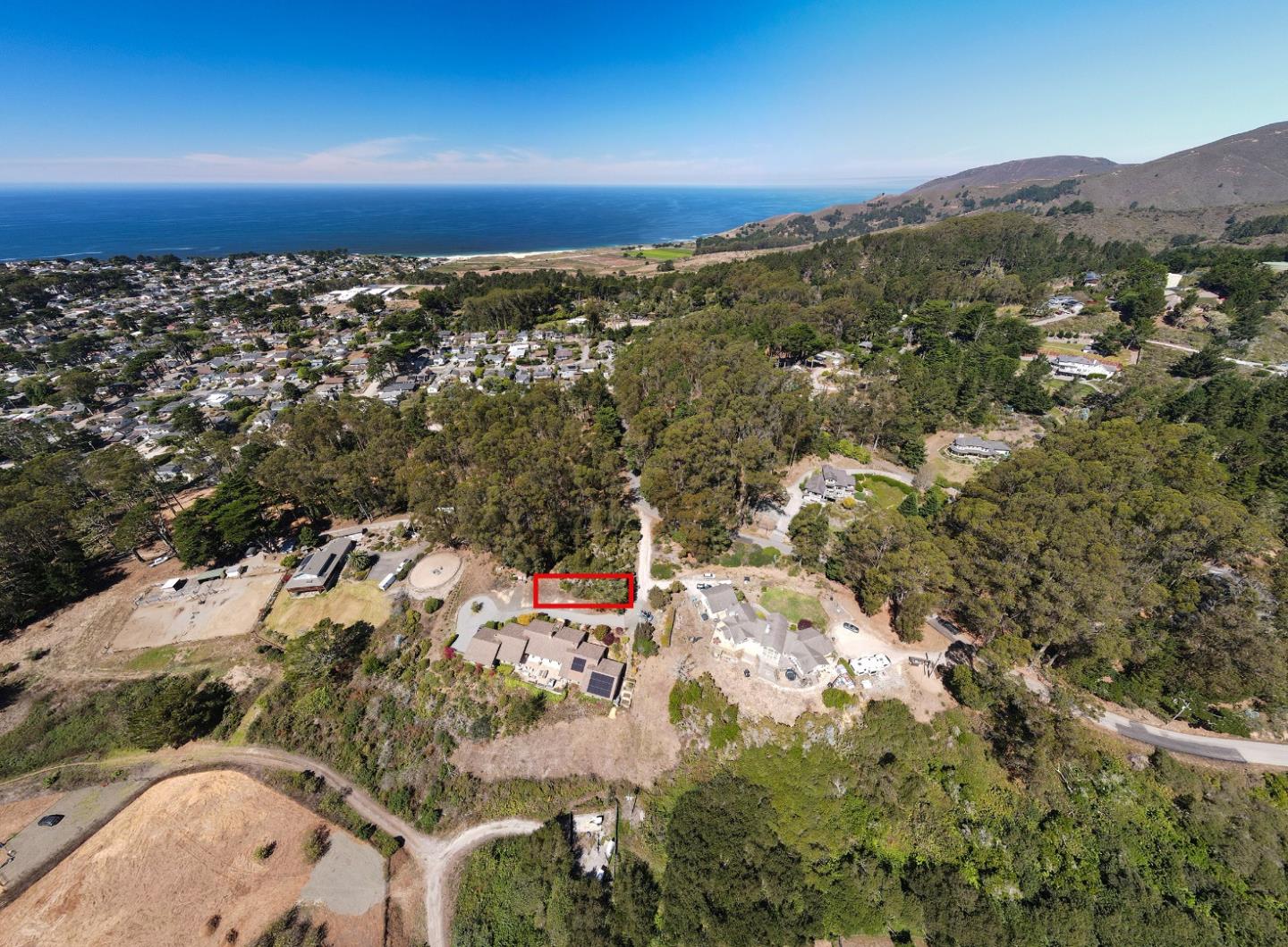 Vacant lot outlined in red amid wooded homes, with the Pacific Ocean and coastal hills beyond.