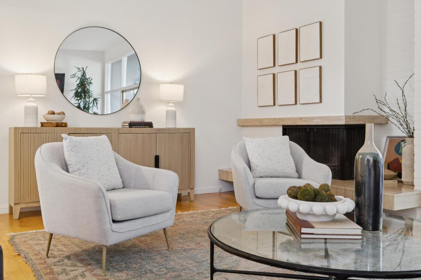 Staged living room with two light armchairs, a fireplace, and a round mirror above a wood sideboard.