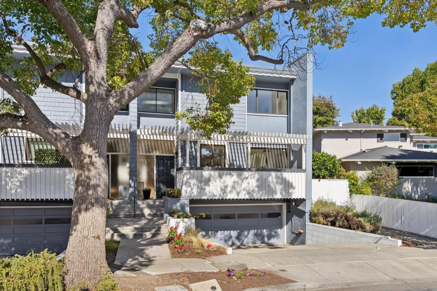 Two-story gray home with white balcony railings, two garages, and a large tree in front.