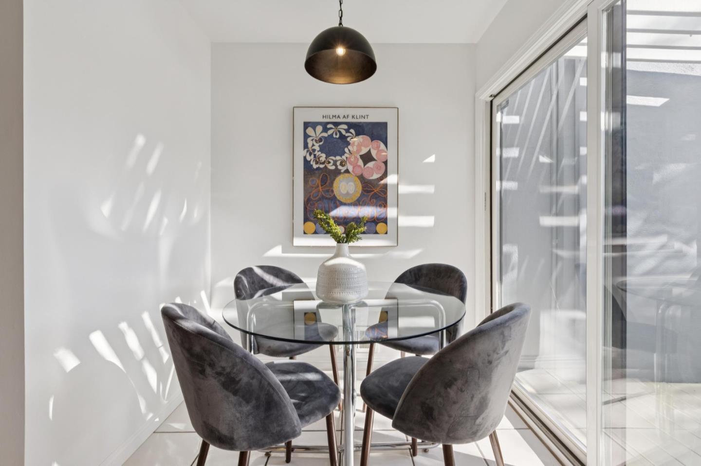 Dining area with a round glass table, gray chairs, and framed artwork beside sliding glass doors.