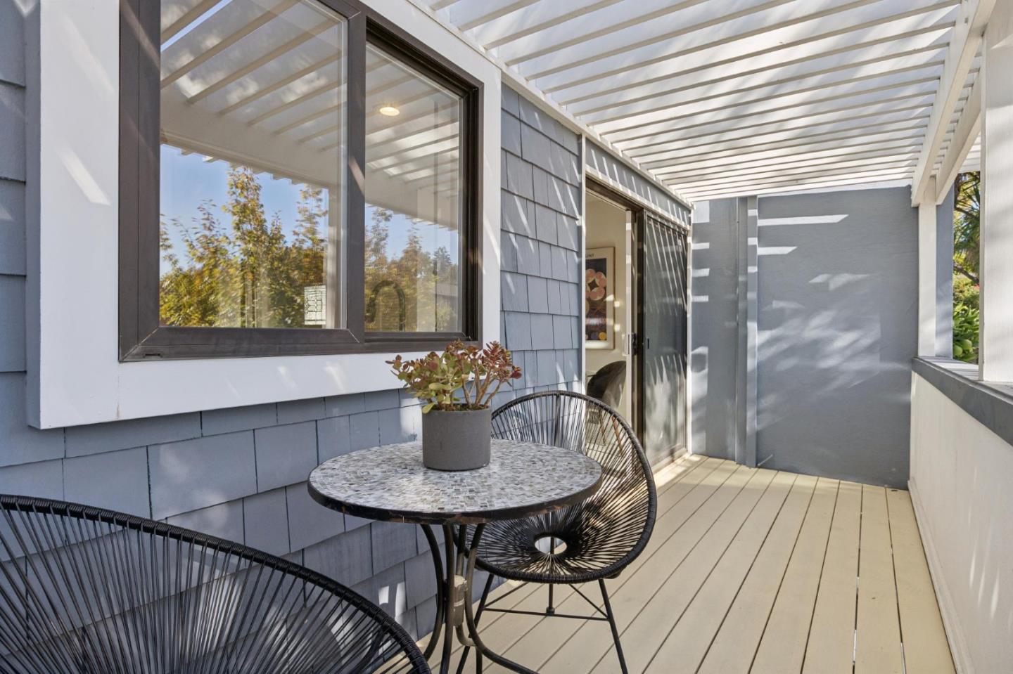 Covered deck with a white pergola roof, round table, wire chairs, and potted plant.
