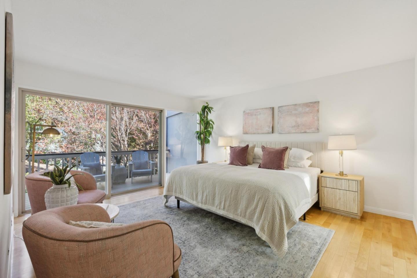 Staged bedroom with a king bed, blush lounge chairs, and sliding doors to a tree-lined balcony.