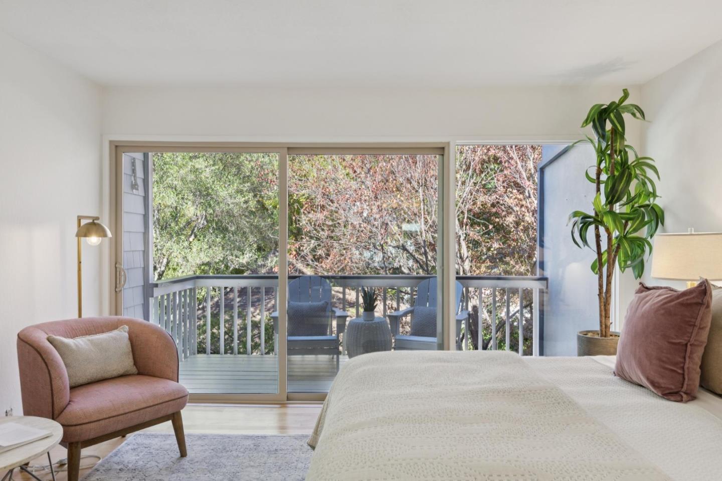 Bedroom with sliding doors opening onto a leafy balcony, furnished with a bed and blush armchair.