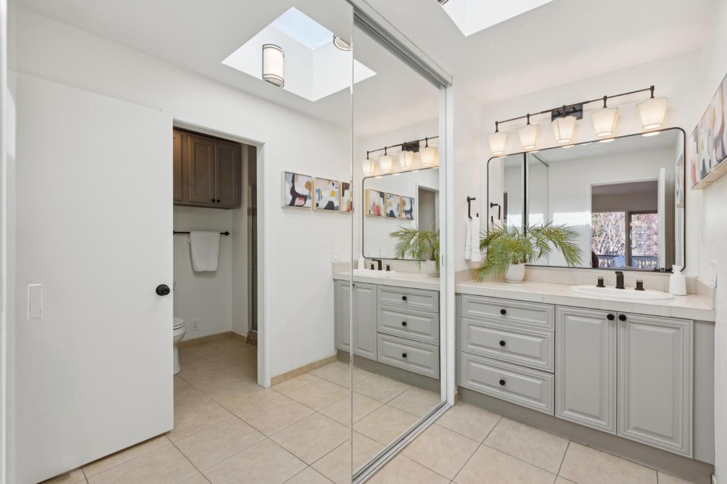 Bright bathroom with a white double vanity, large mirrors, tiled floor, and skylight.